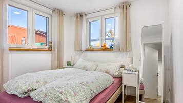 Un dormitorio con cama, una mesita de noche con lámpara, un espejo y una ventana con cortinas.
