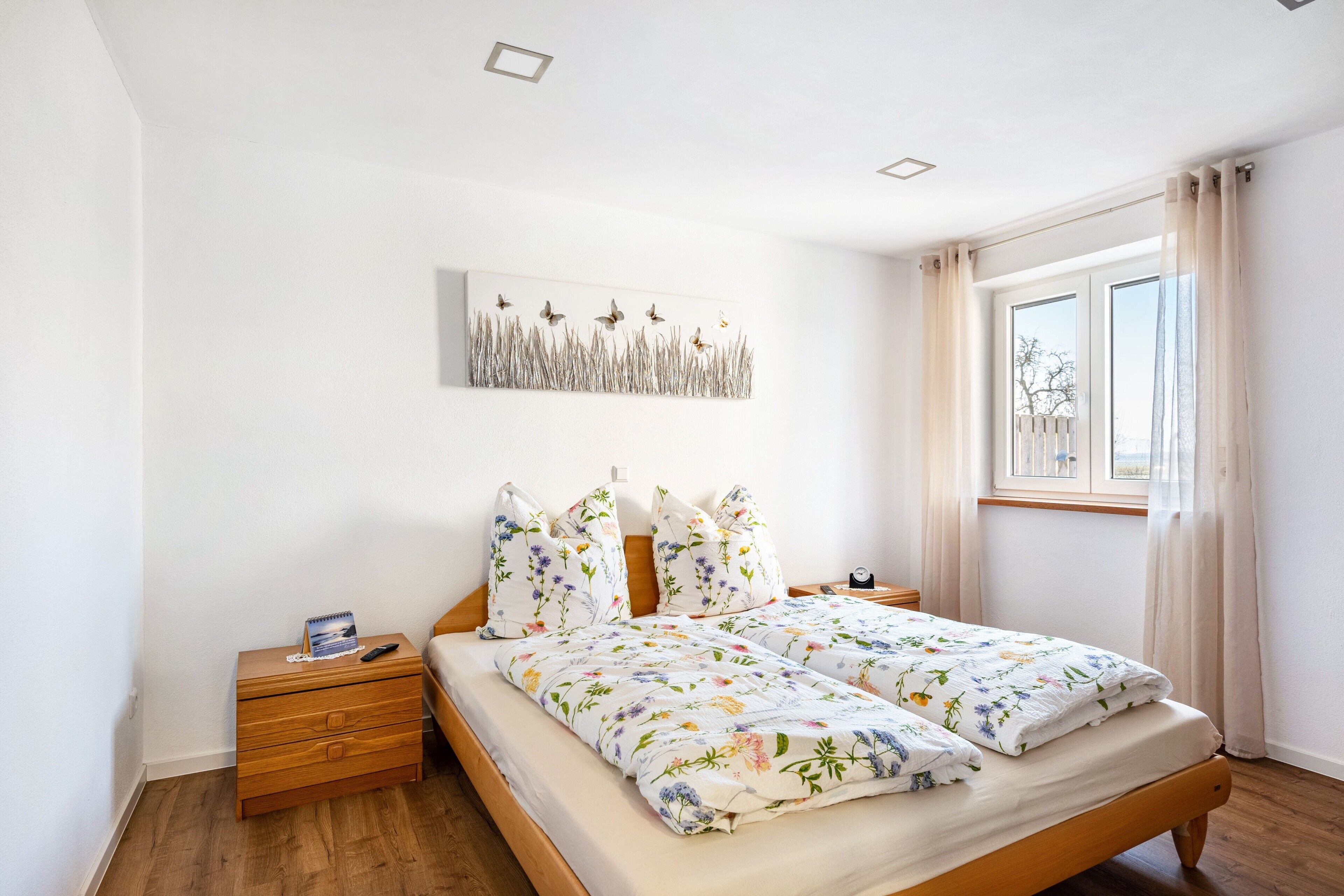Une chambre à coucher avec un lit en bois, une table de chevet, une œuvre d’art accrochée au mur et une fenêtre avec des rideaux.