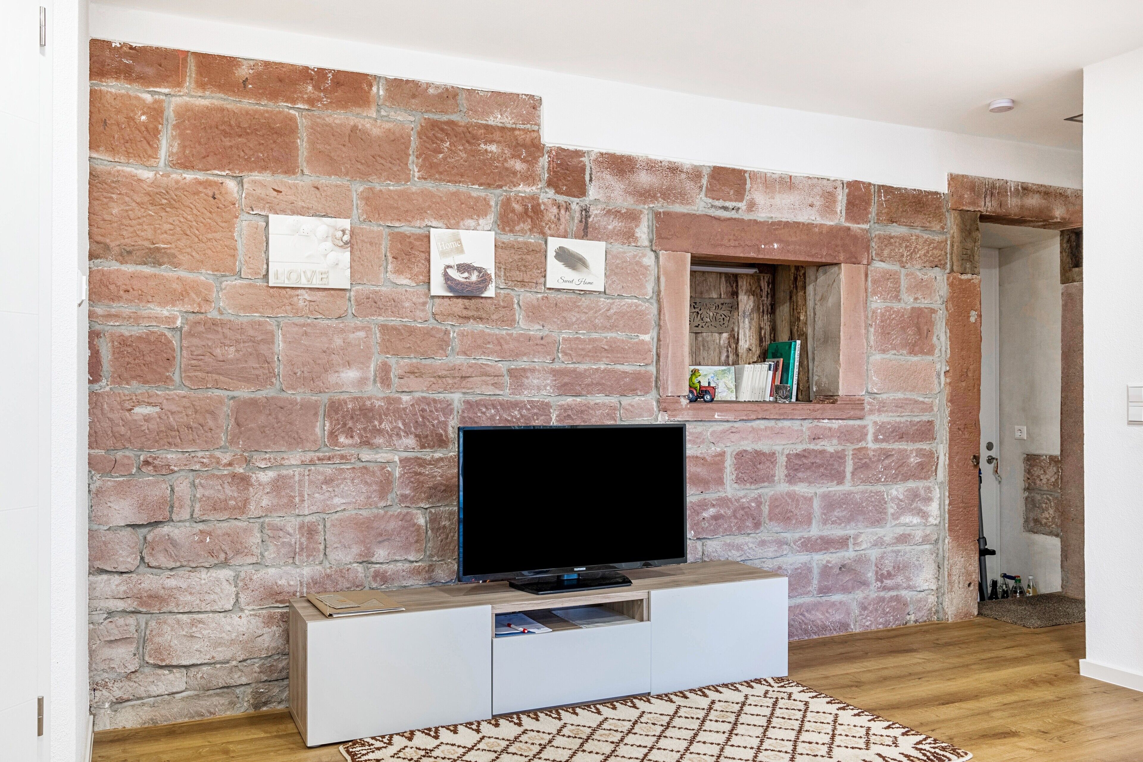 Una stanza con un muro in mattoni, una TV a schermo piatto su un mobile bianco, una piccola finestra con uno scaffale e un tappeto decorato.