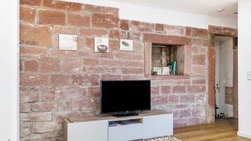 Una stanza con un muro in mattoni, una TV a schermo piatto su un mobile bianco, una piccola finestra con uno scaffale e un tappeto decorato.