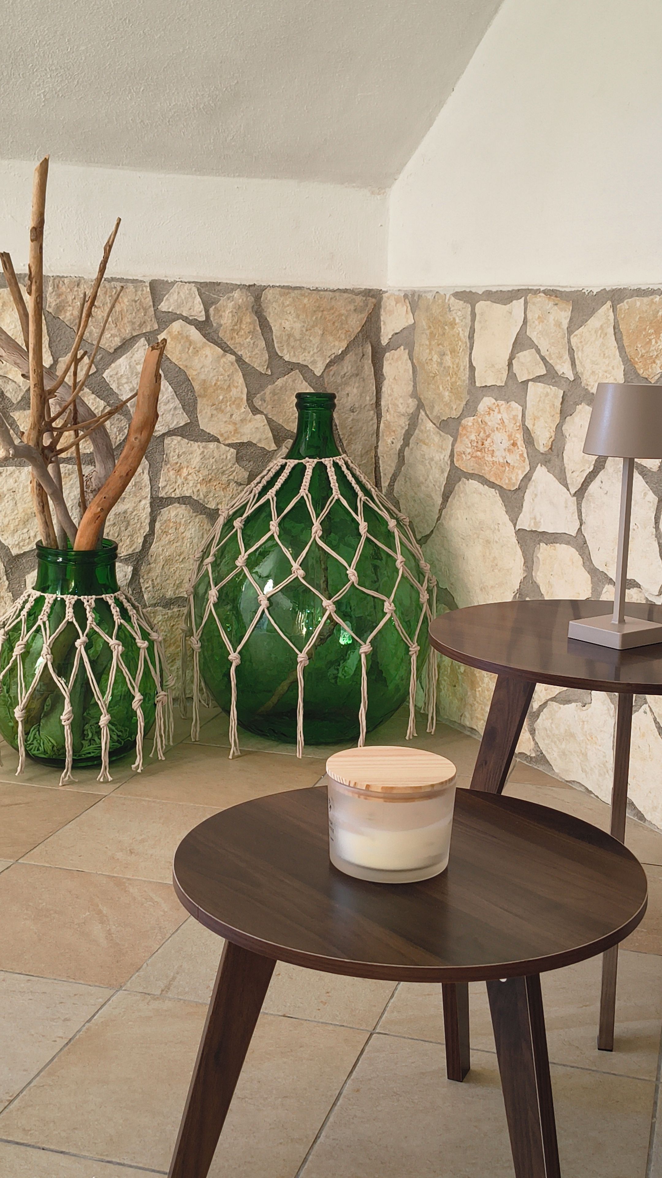 A room with a stone wall, a round wooden table, and two green glass vases with macrame decorations.