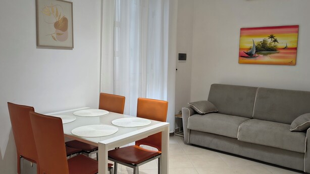 A dining area with a table and chairs, a sofa, a painting, and a nightstand with a lamp.