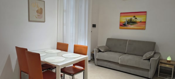 A dining area with a table and chairs, a sofa, a painting, and a nightstand with a lamp.