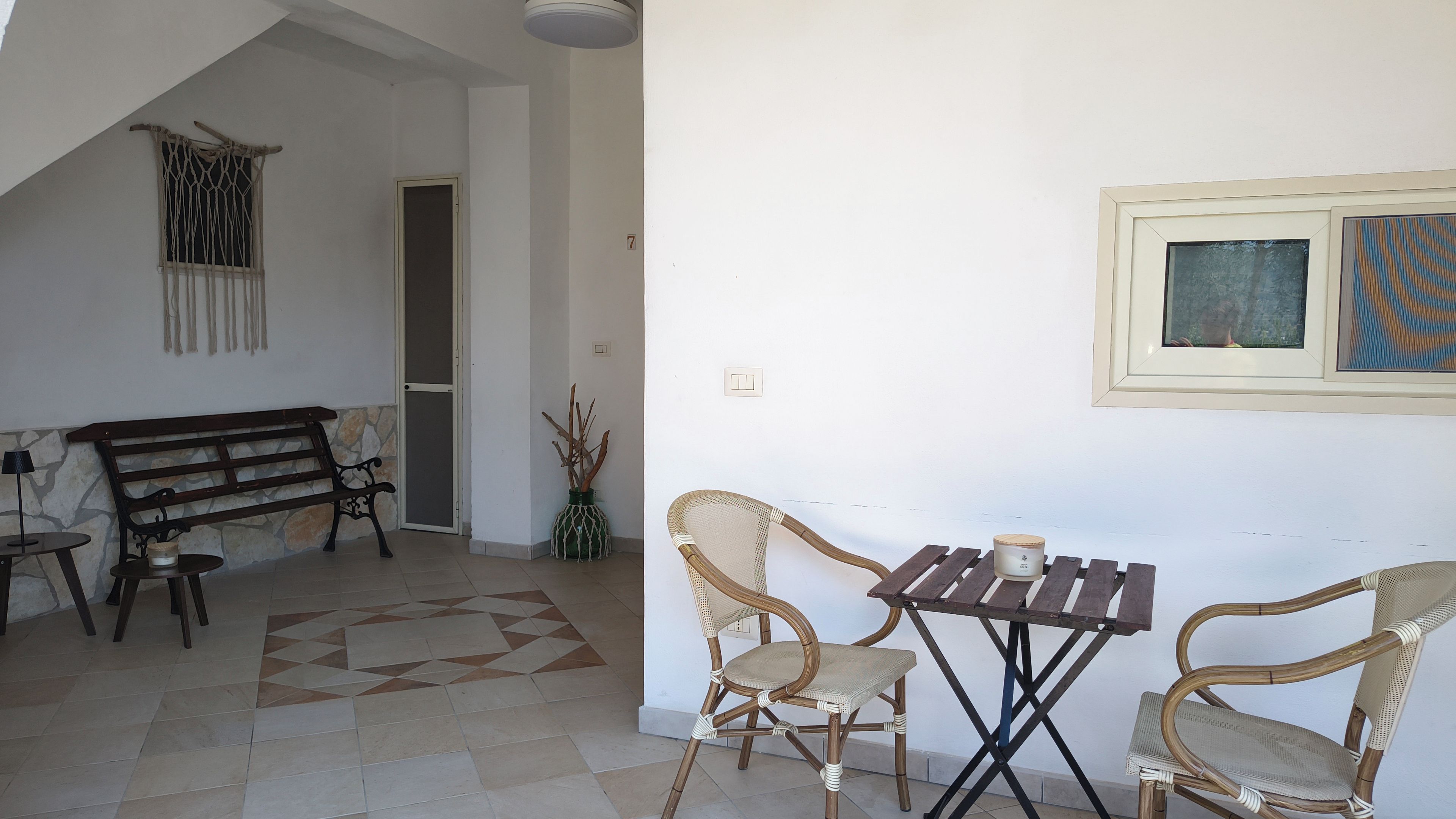 A room with a wooden bench, two wicker chairs, a small table, a potted plant, and a door with a number.