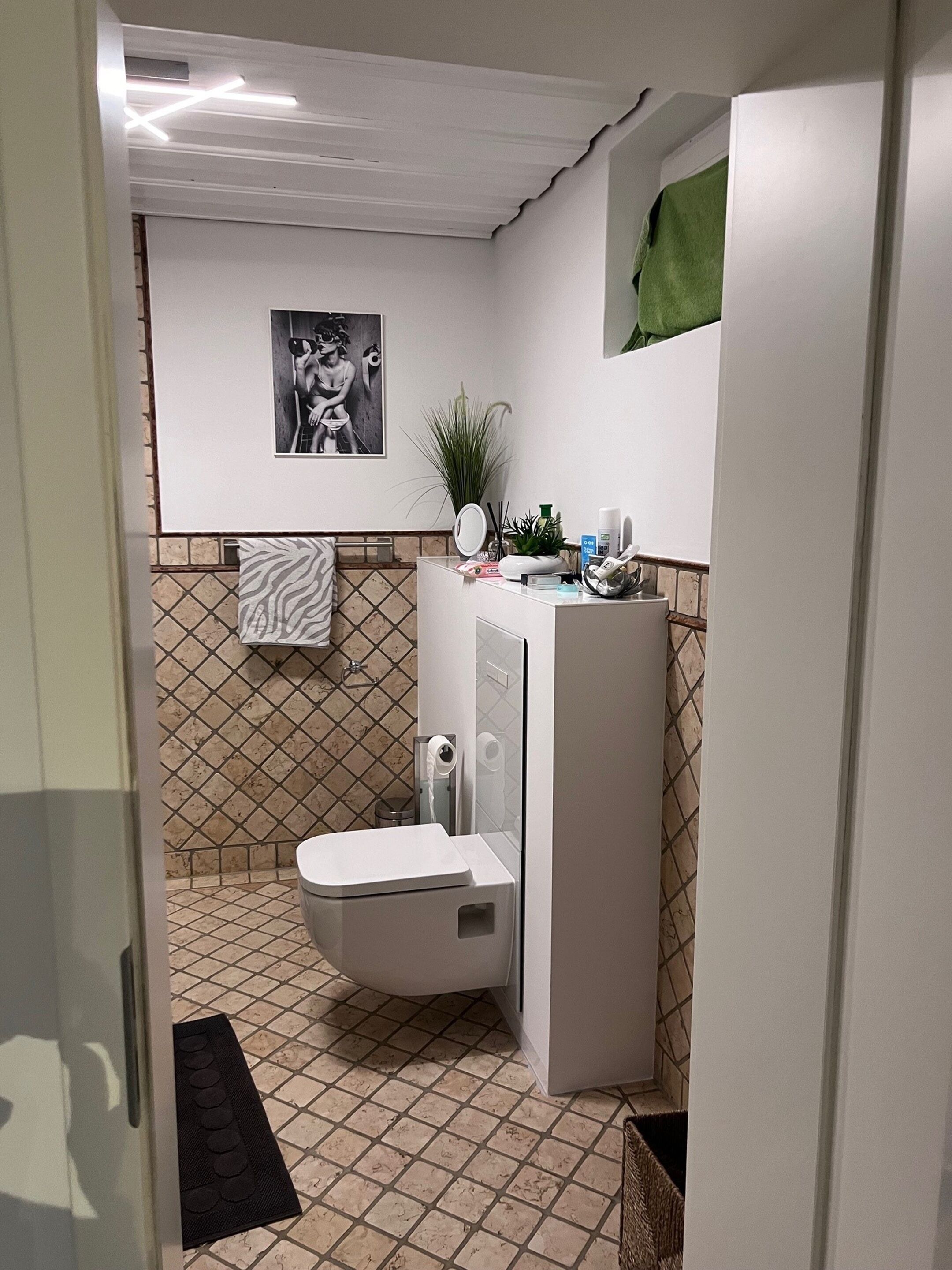 A compact bathroom with a white toilet, a small sink, and a mirror. There is a green towel hanging on the wall and a picture frame above the toilet.