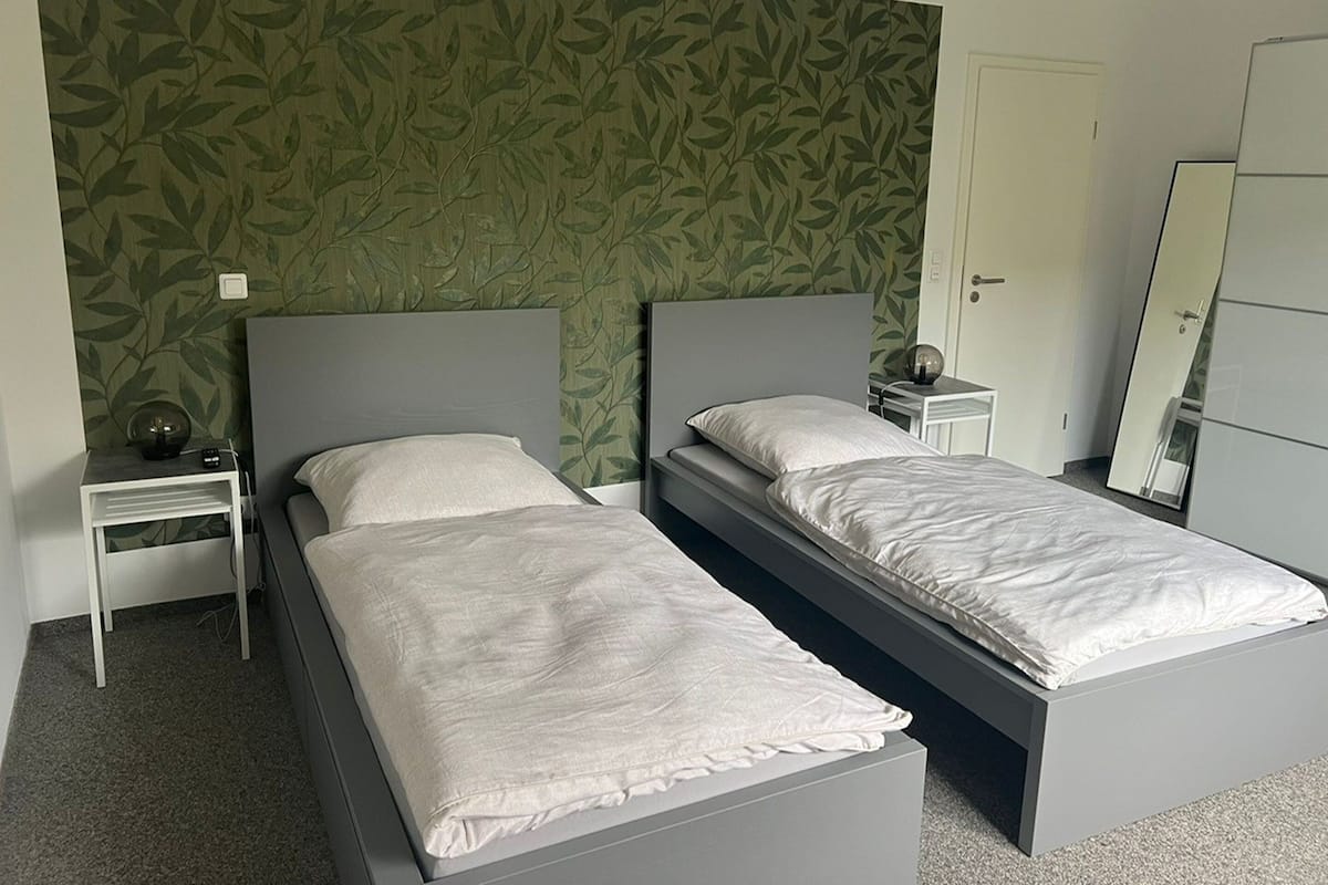 Een tweepersoonsbed met een grijs hoofdbord en witte beddengoed, een nachtkastje met een lamp, en een kledingkast met een spiegel.