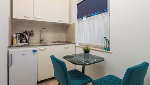 A compact kitchen with white cabinets, a built-in refrigerator, a sink, and a window with a white curtain. There is a small table with a potted plant and teal chairs. - Villa Chiara Opatija Apartment Moderna (Ičići)