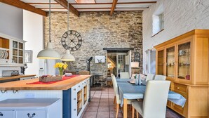 A rustic kitchen with a stone wall, wooden cabinets, and a dining area with a table and chairs. - The Old Farmhouse (Looe)