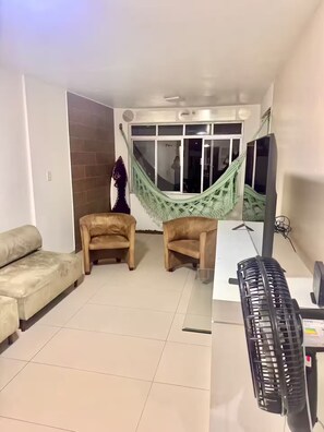 Ein Wohnzimmer mit einem beigefarbenen Sofa, zwei Sesseln, einer grünen Hängematte, einem Fernseher und einem schwarzen Heizgerät.