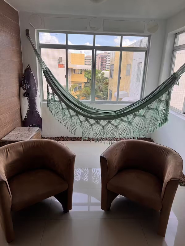 Ein Zimmer mit zwei braunen Ledersesseln, einer Hängematte und Blick auf Gebäude durch große Fenster.