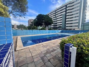 Ein Schwimmbecken mit Steinfliesen, umgeben von einem blauen Zaun, neben einem modernen, mehrstöckigen Gebäude.