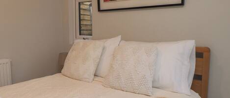 Una cama bien tendida con una colcha blanca y dos almohadas, una obra enmarcada encima de la cama y una ventana con persianas a medio bajar.