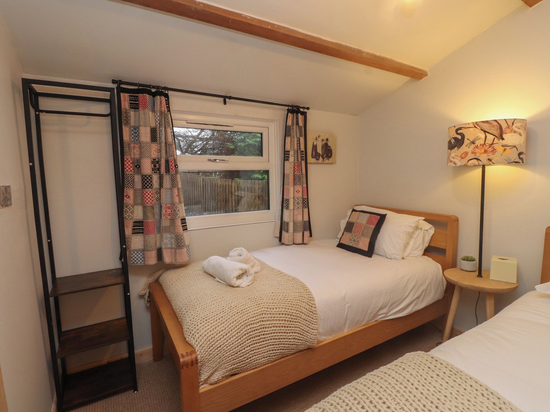 A cozy bedroom with a wooden bed, a window with curtains, a bedside table with a lamp, and a small shelf.