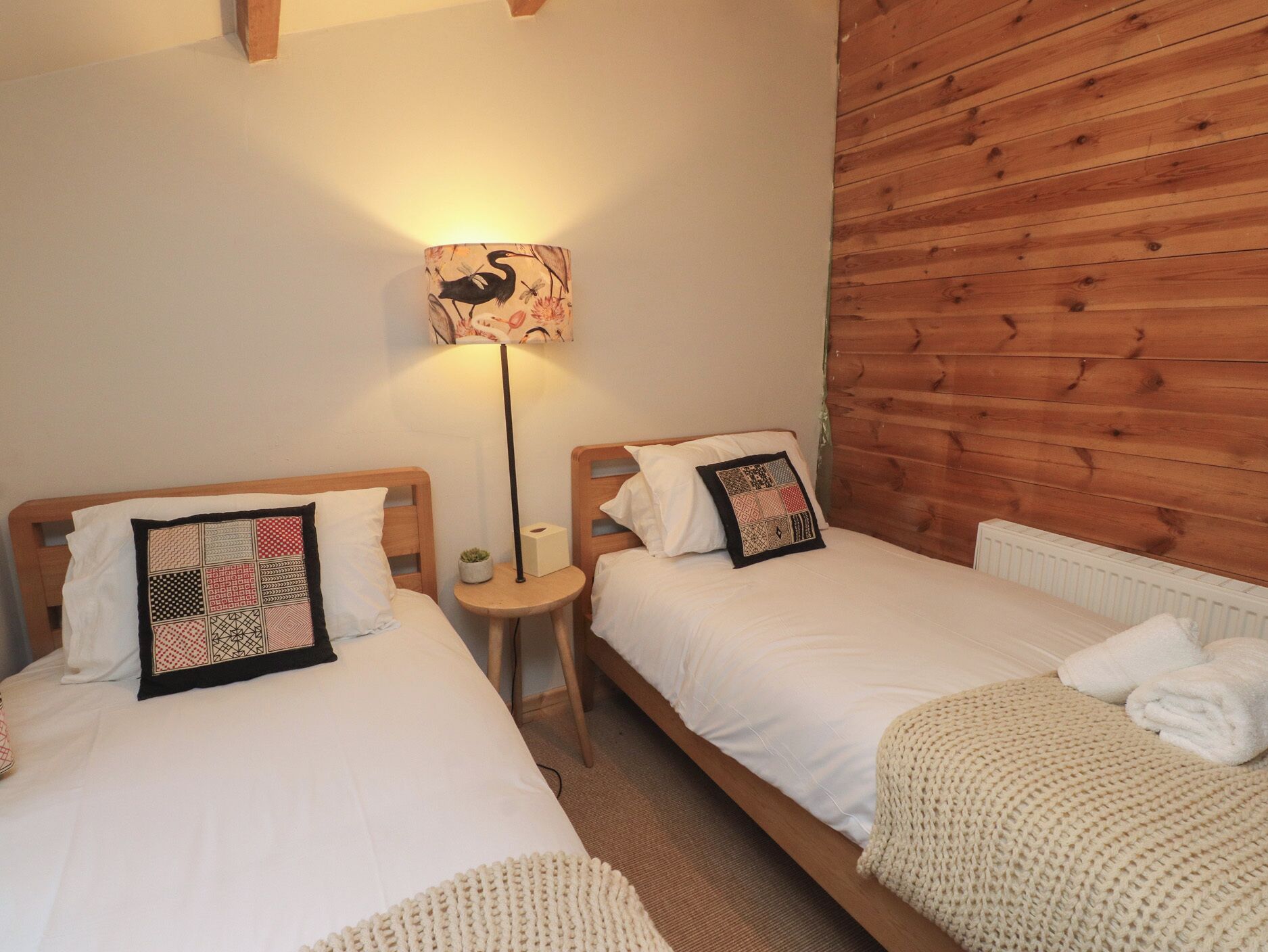 A room with two single beds, each with a white duvet and a patterned pillow. There is a small round table with a lamp between the beds. The room has wooden paneling on one wall and a radiator under the beds.