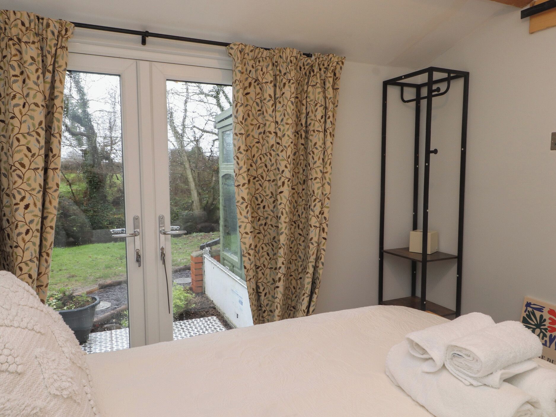A bedroom with a bed, a window with curtains, a small shelf, and a view of the outdoors.