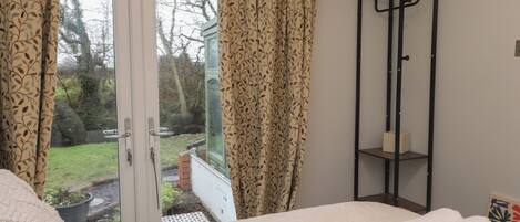 A bedroom with a bed, a window with curtains, a small shelf, and a view of the outdoors.