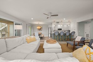 A modern living room with a white sofa, a dining area with bar stools, and a kitchen in the background. - Surfside 12A2 | Luxury Beachview Living on Sanibel (Sanibel)