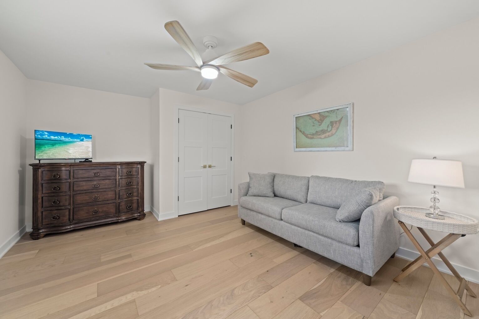 A room with a wooden floor, a grey sofa, a wooden cabinet, a ceiling fan, a white door, and a small round table with a lamp.