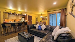 A living room with a leather sofa, ottoman, and bar stools. There is a kitchen area with wooden cabinets and a dining table. - New Listing: Pool, Hot Tub, Free shuttle (Keystone)