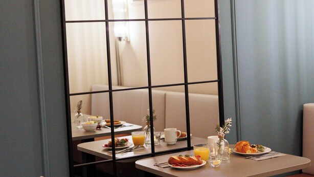 Uma área de refeições com uma cadeira branca de encosto alto, uma mesa com itens de café da manhã e um espelho grande refletindo o ambiente.