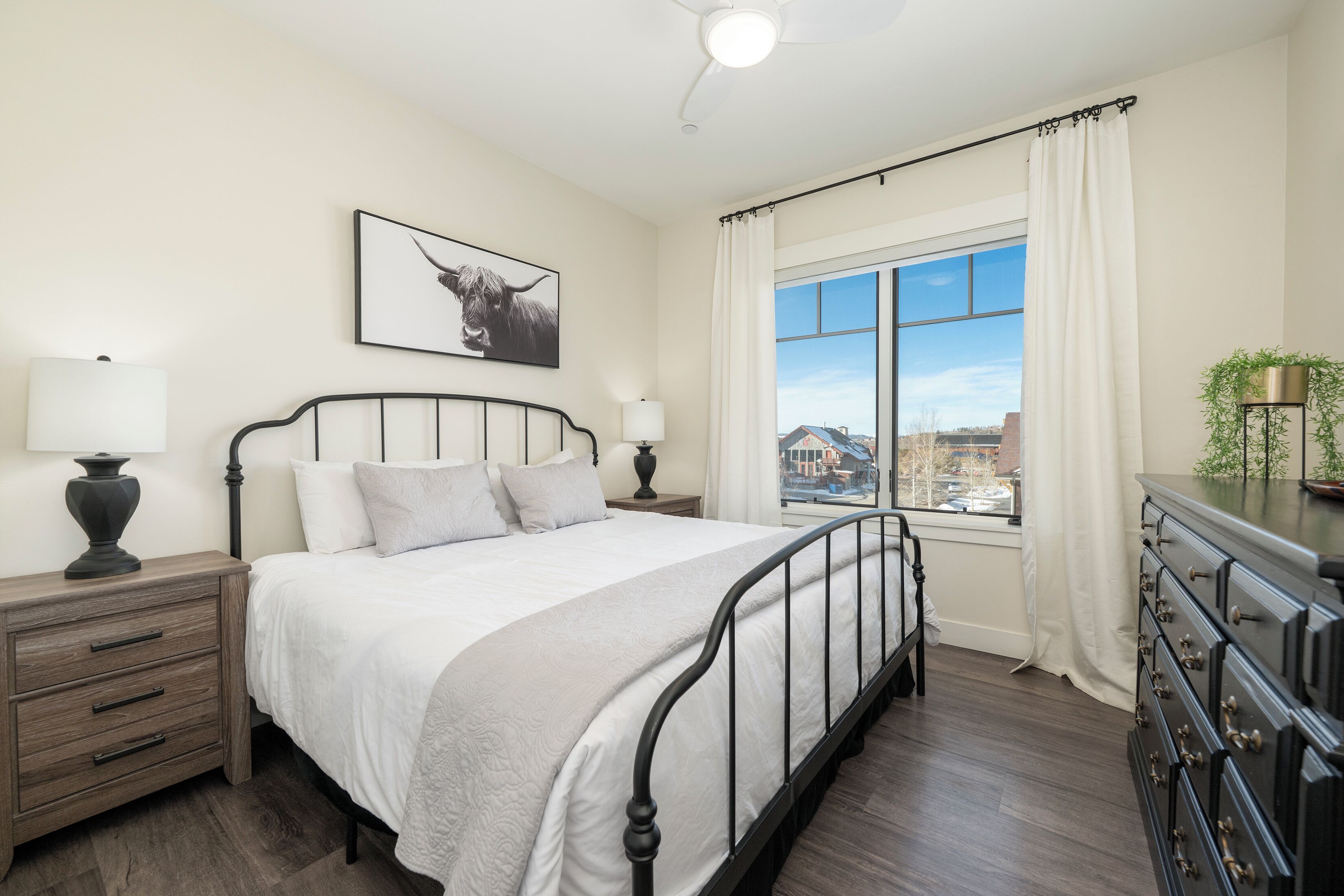 A bedroom with a bed, bedside tables, a dresser, a window with curtains, and a city view.