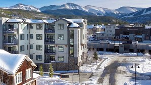 Un immeuble à plusieurs étages, dont le toit est recouvert de neige, avec un espace de stationnement et une chaîne de montagnes en arrière-plan.