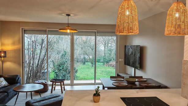 Ein modernes Wohnzimmer mit Sofa, Couchtisch und Essbereich. Ein großes Fenster bietet Blick auf Bäume und eine Rasenfläche. Zwei Pendelleuchten hängen von der Decke.