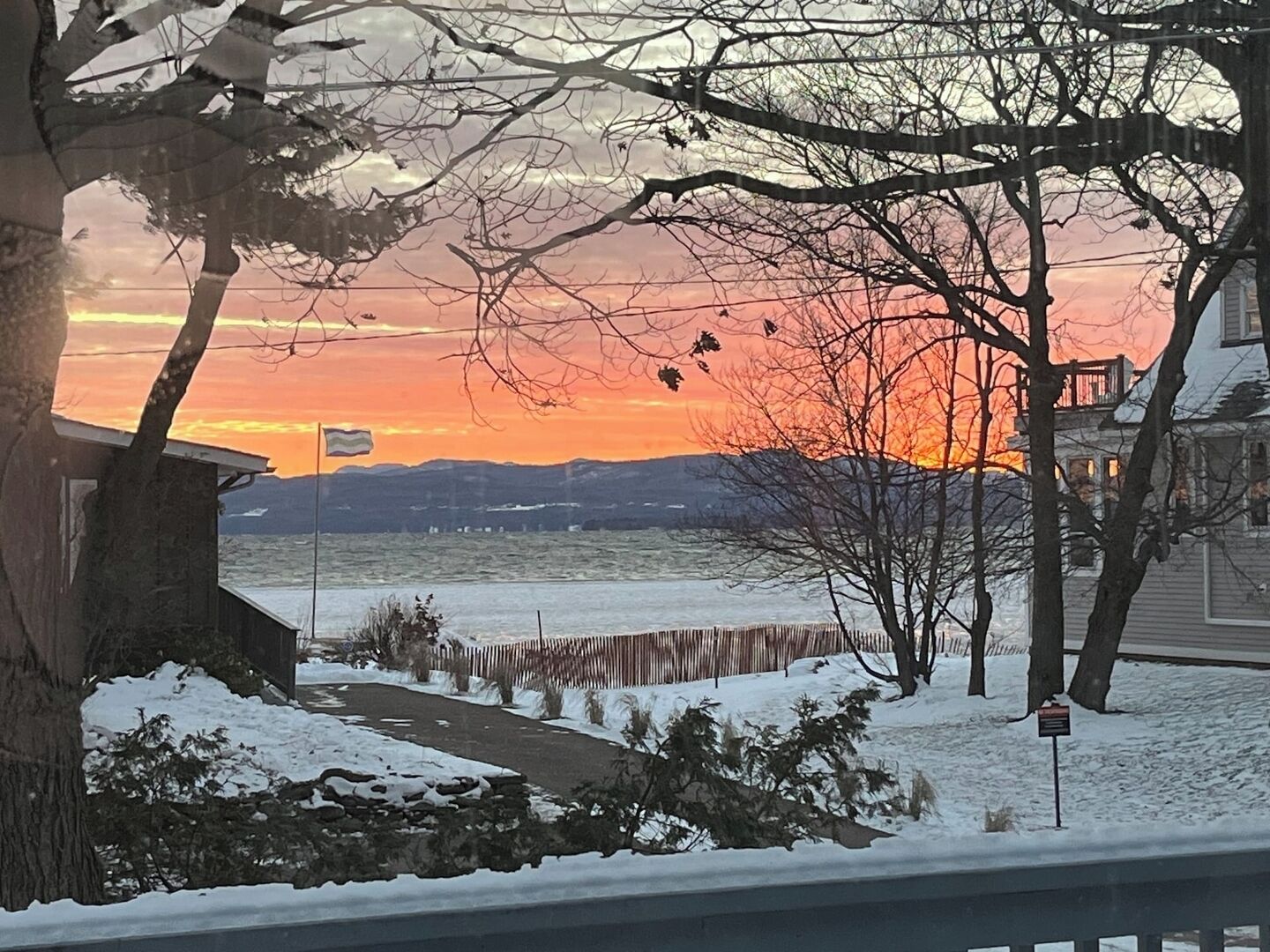 A snowy landscape with a sunset over a body of water, trees, and a house.