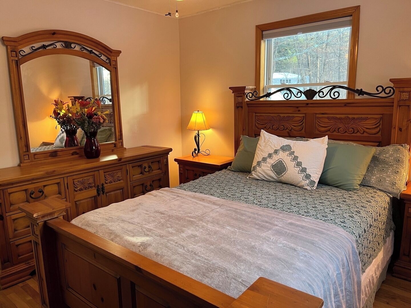 Een slaapkamer met een houten bed, een nachtkastje met een lamp, een kaptafel met spiegel en een raam met uitzicht op bomen.
