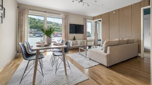 A modern living room with a sofa, dining table, and chairs. There is a flat-screen TV mounted on the wall and a large window with curtains.