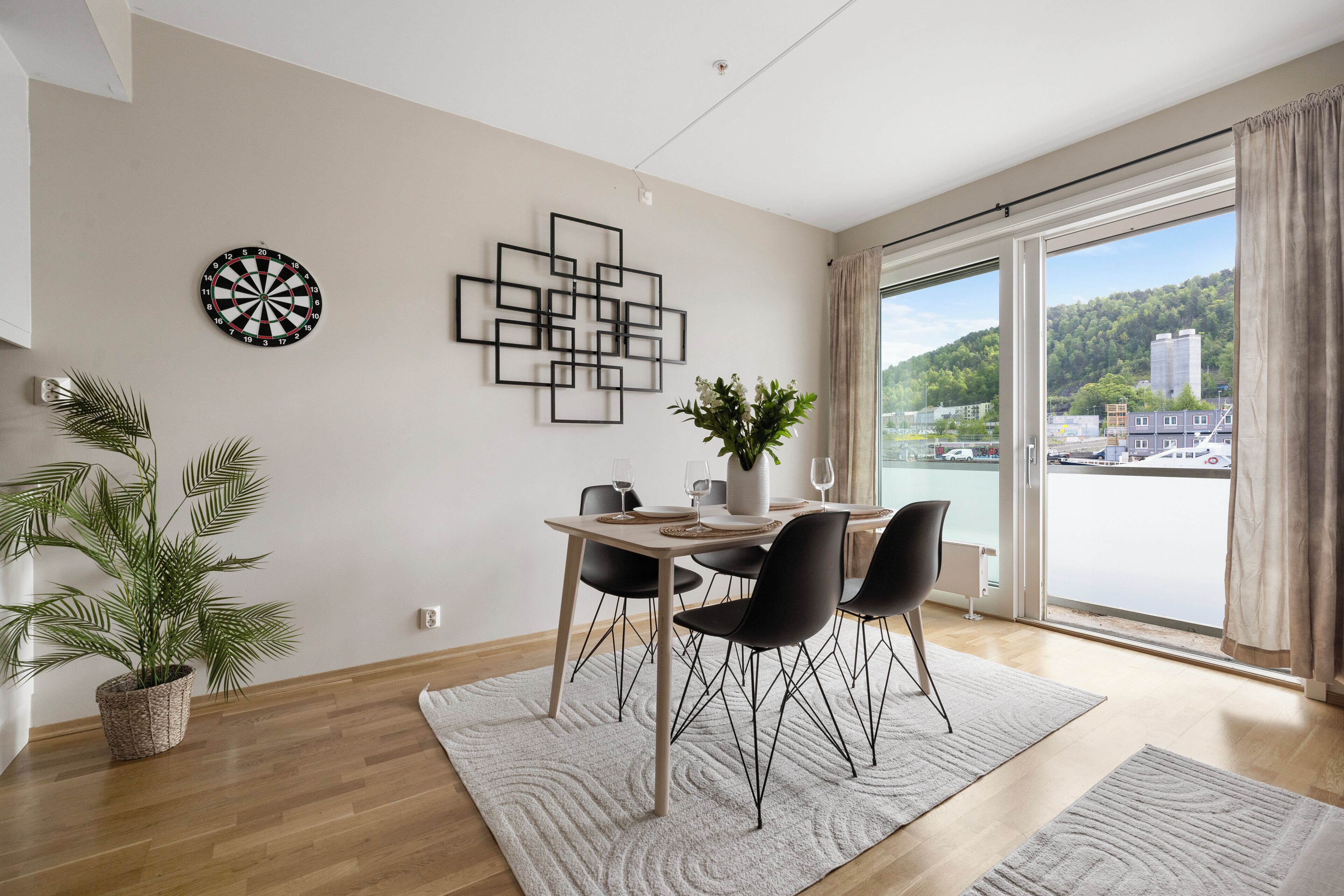 A dining area with a table set for four, a potted plant, a dartboard, and a modern wall art piece.