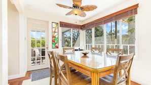 A dining room with a glass table, wooden chairs, a ceiling fan, and large windows with a view of greenery. - Hale Ho‘omalie- Peaceful Mānoa Valley Retreat near UH (Honolulu)