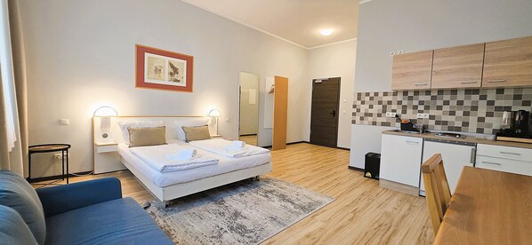 Une chambre d’hôtel moderne comprenant un lit, des tables de chevet, une petite table, un canapé, un coin cuisine avec des placards et un plan de travail, ainsi qu’un tableau encadré au mur.