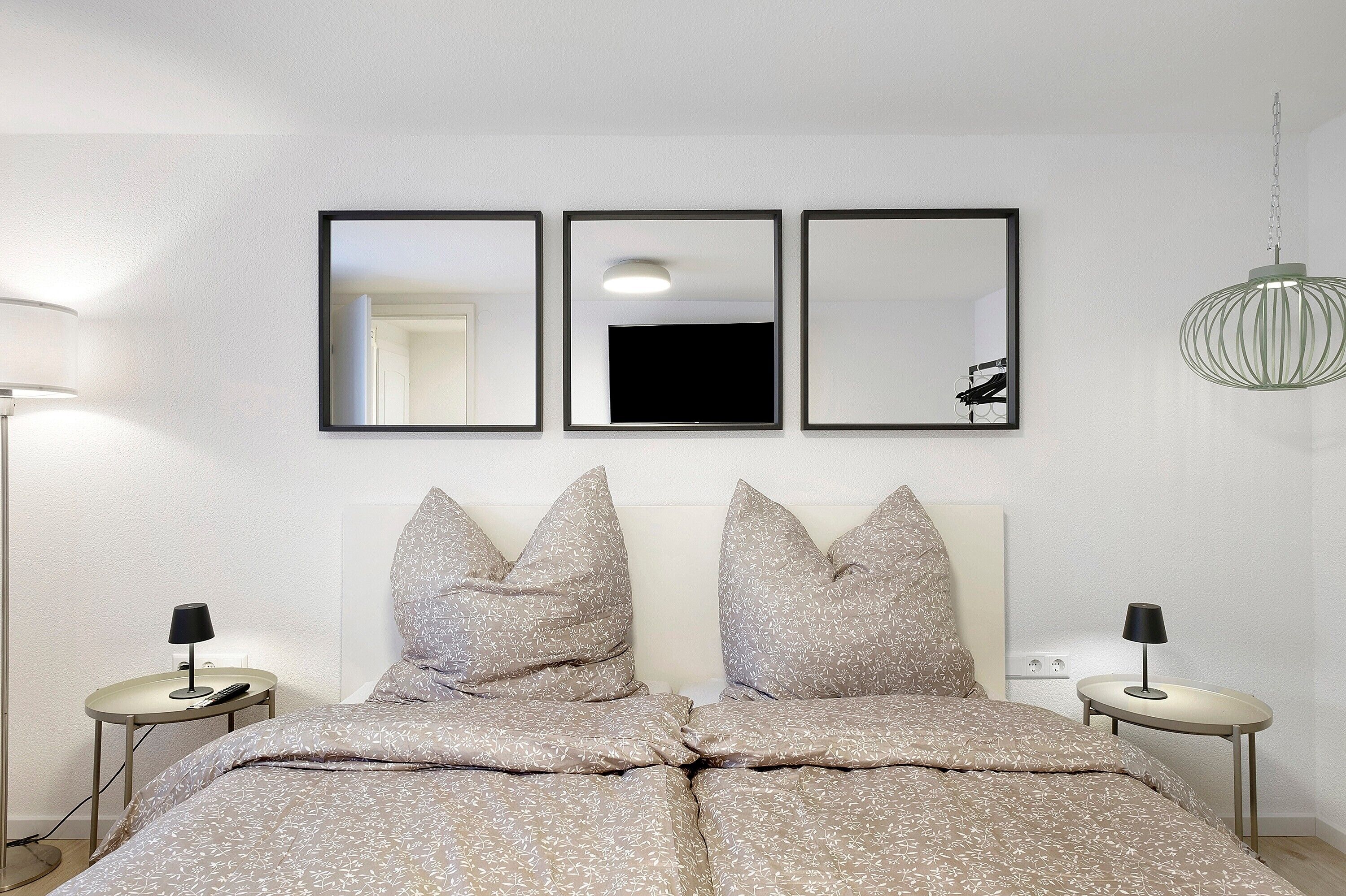 Un dormitorio con cama, dos mesitas de noche, una lámpara, un televisor y tres espejos enmarcados en la pared.