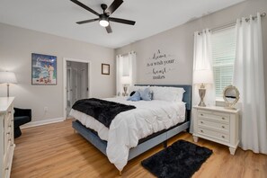 A bedroom with a bed, a ceiling fan, a wall with a motivational quote, a framed picture, a bedside table with a lamp, and a window with curtains. - Magic & Muggles: Private Pool/Spa, BBQ, Game Room! (Kissimmee)