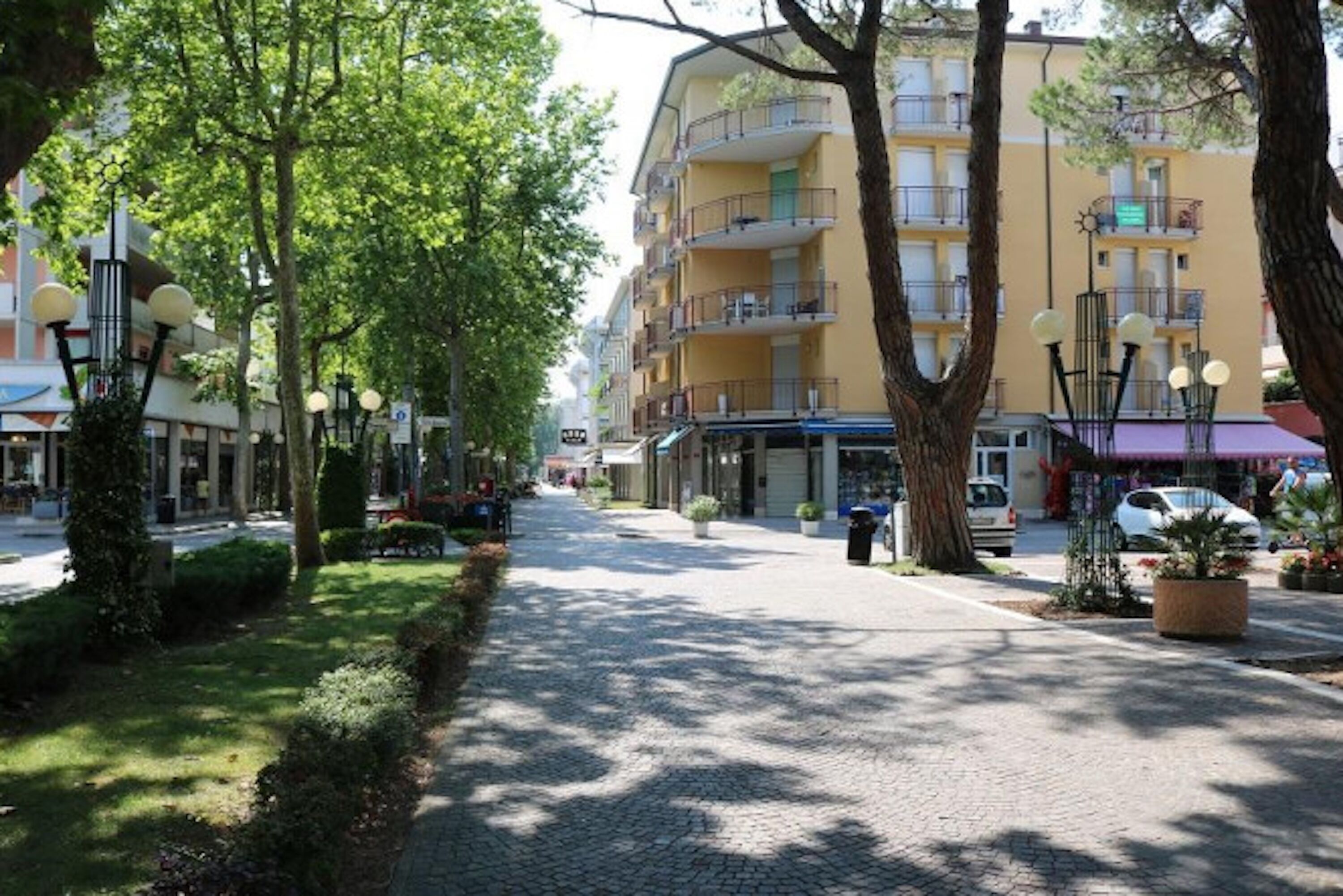 A tree-lined cobblestone street with multi-story buildings, including a yellow building with balconies.