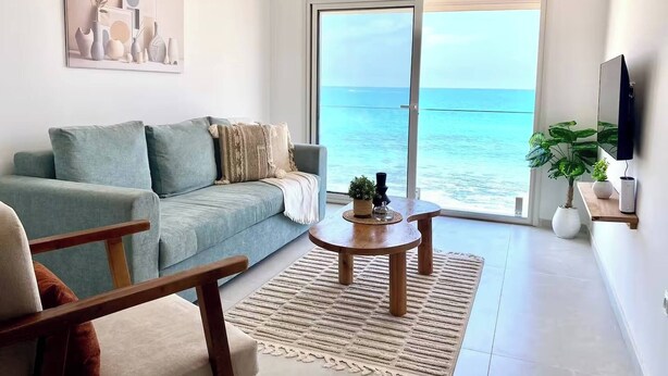 A living room with a sofa, coffee table, and a view of the ocean through large sliding doors. - L'écume Blanche - Apartment With Feet in the Water (La Marsa)