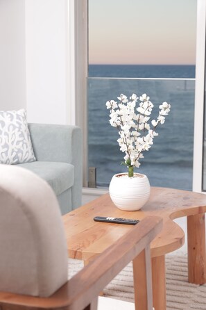 A room with a view of the sea, a white vase with flowers on a wooden table, and a light blue sofa.