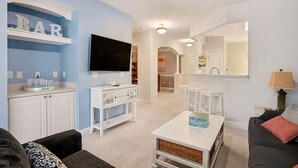 Una sala de estar con televisor, muebles blancos, una zona de cocina y un letrero de bar en la pared.
