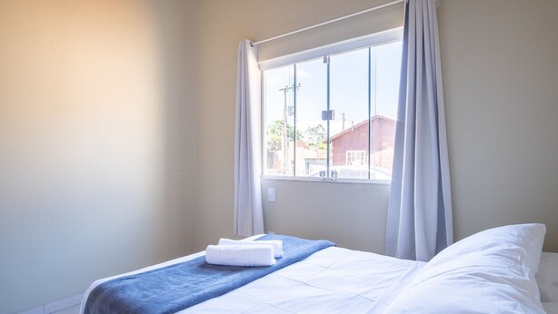 A neatly made bed with white linens and a blue blanket, a window with white curtains, and a view of buildings outside. - 2-bedroom House With Barbecue Area Wil0012 (Imbituba)