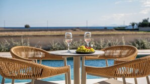 Une table placée au bord de la piscine, avec deux verres à vin et une assiette de raisins, entourée de chaises en osier, donnant sur une étendue d’eau.