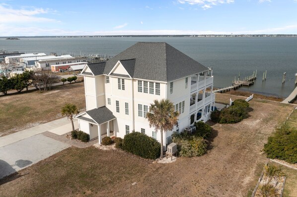 A two-story house with a dock extending into a body of water, surrounded by a grassy area and some trees. - Soundfront Living with Ocean Views, Private Dock & Pool (Emerald Isle)