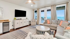 A spacious living room with a large flat-screen TV, a ceiling fan, and a view of the ocean through large windows. - Gulf-Front Duplex | Pet Friendly | Shared Pool (Gulf Shores)