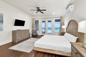 Ein Schlafzimmer mit Bett, Fernseher, Deckenventilator, Klimaanlage, einem Holzschrank, einem Sessel und Meerblick durch eine Schiebetür.