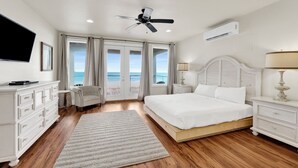 Un dormitorio con cama, televisor, ventilador de techo, aire acondicionado, mesita de noche, lámpara, sillón y vistas al océano a través de amplias ventanas.