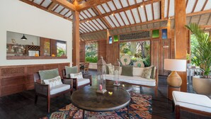 A spacious lounge area with a high ceiling, wooden beams, and a large window offering a view of lush greenery. There are multiple seating options including sofas and chairs, and a round table with a decorative centerpiece. - Private luxury 2 bedroom villa in the heart of Berawa! (Canggu)
