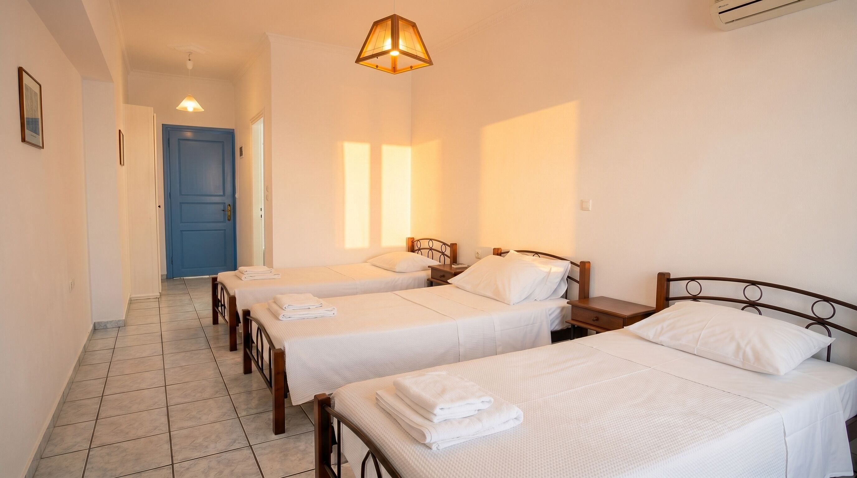 Habitación de hotel con tres camas individuales, una puerta azul, un pasillo y una lámpara colgante.