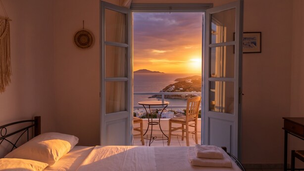 A bedroom with a bed, a chair, a table, and an open window with a view of the sea and a sunset. - Lume D'oro Syros (Kini)