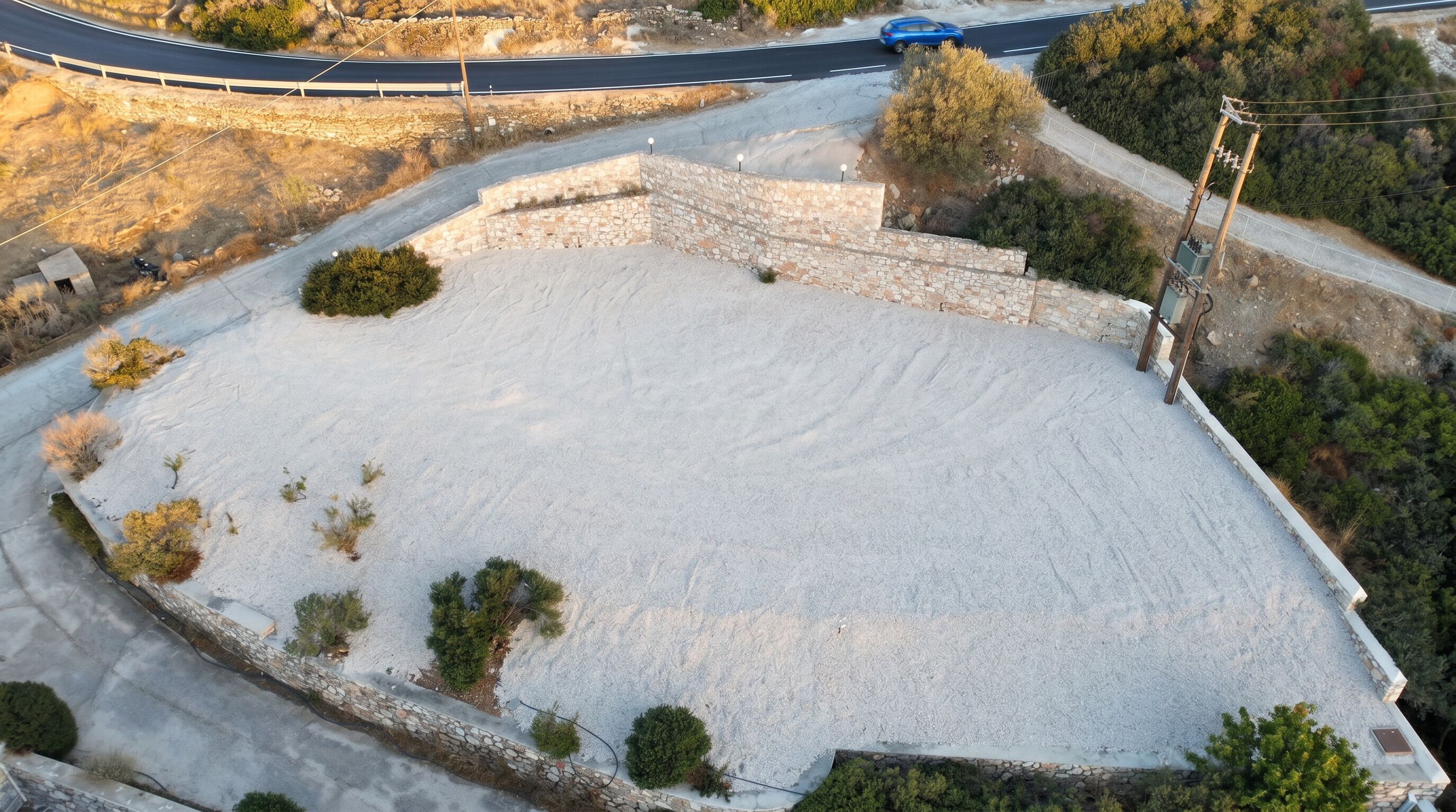 An aerial view of a large, open, paved area with a stone wall and sparse vegetation.