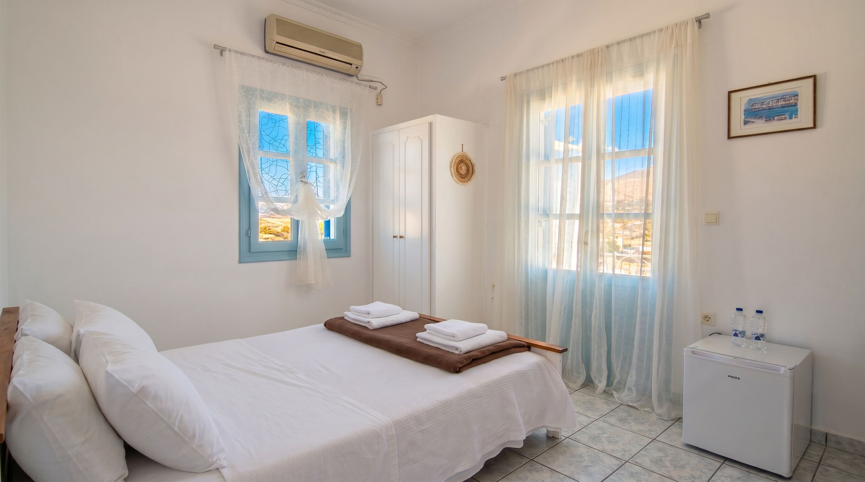 A bedroom with a bed, a bedside table, a refrigerator, and a window with curtains.